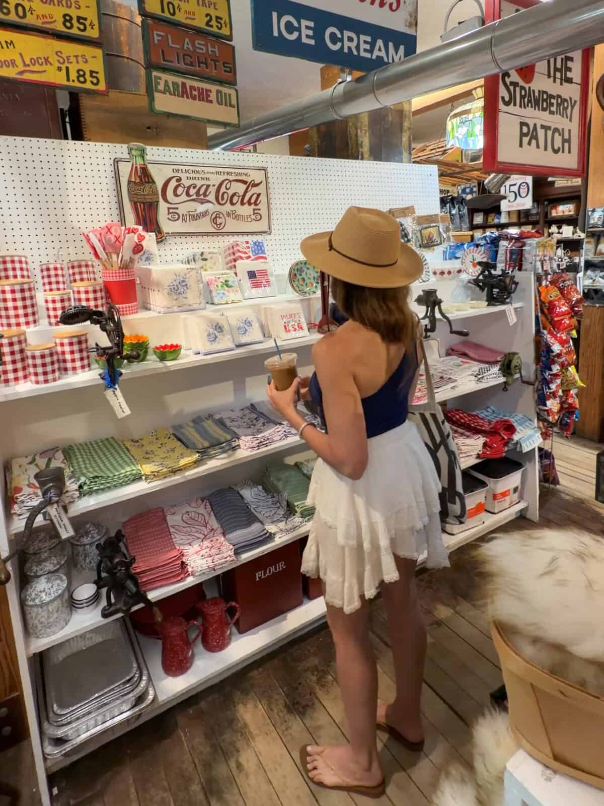 Laura wearing a tan sun hat, a navy tank top, and a white tiered skirt stands on a rustic wooden floor inside The Brewster Store, sipping an iced coffee while browsing shelves filled with vintage-style home goods, textiles, and retro tin signs.