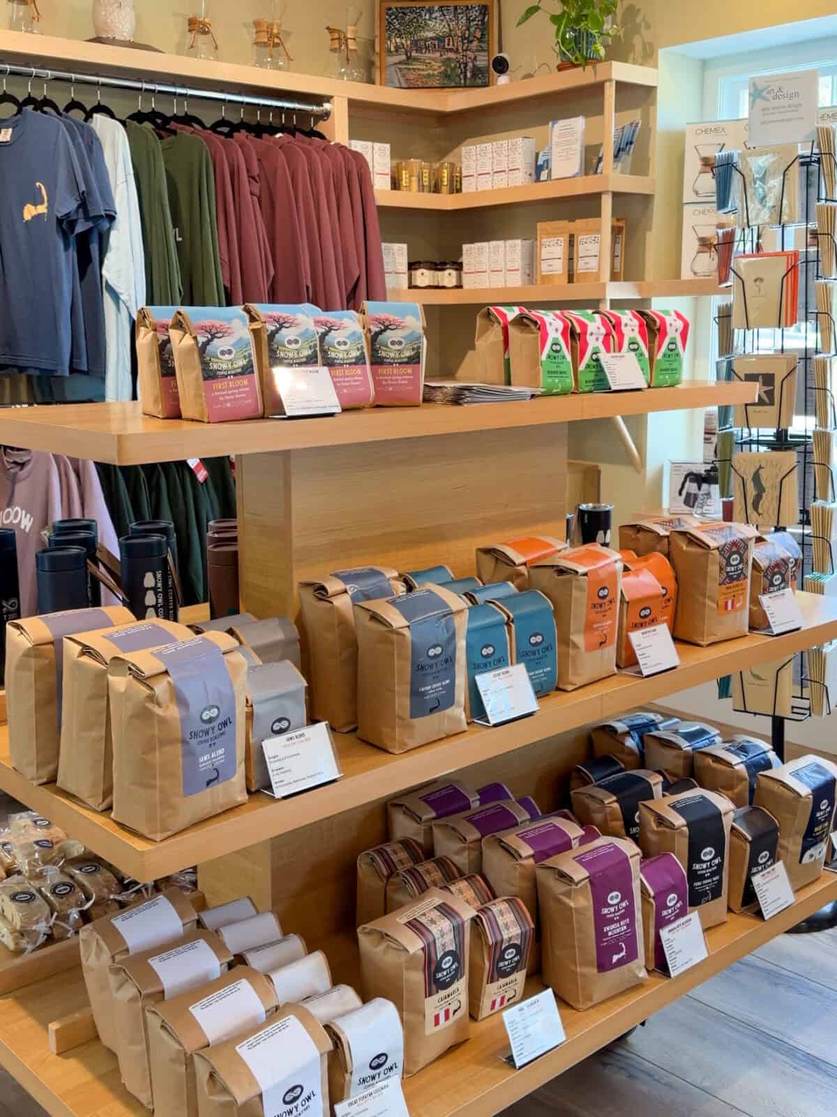 Light wooden display shelves inside the Snowy Owl Coffee Roasters store in Brewster, packed with colorful bags of specialty coffee beans, including "First Bloom" and "Jaws Blend," alongside branded t-shirts and apparel.