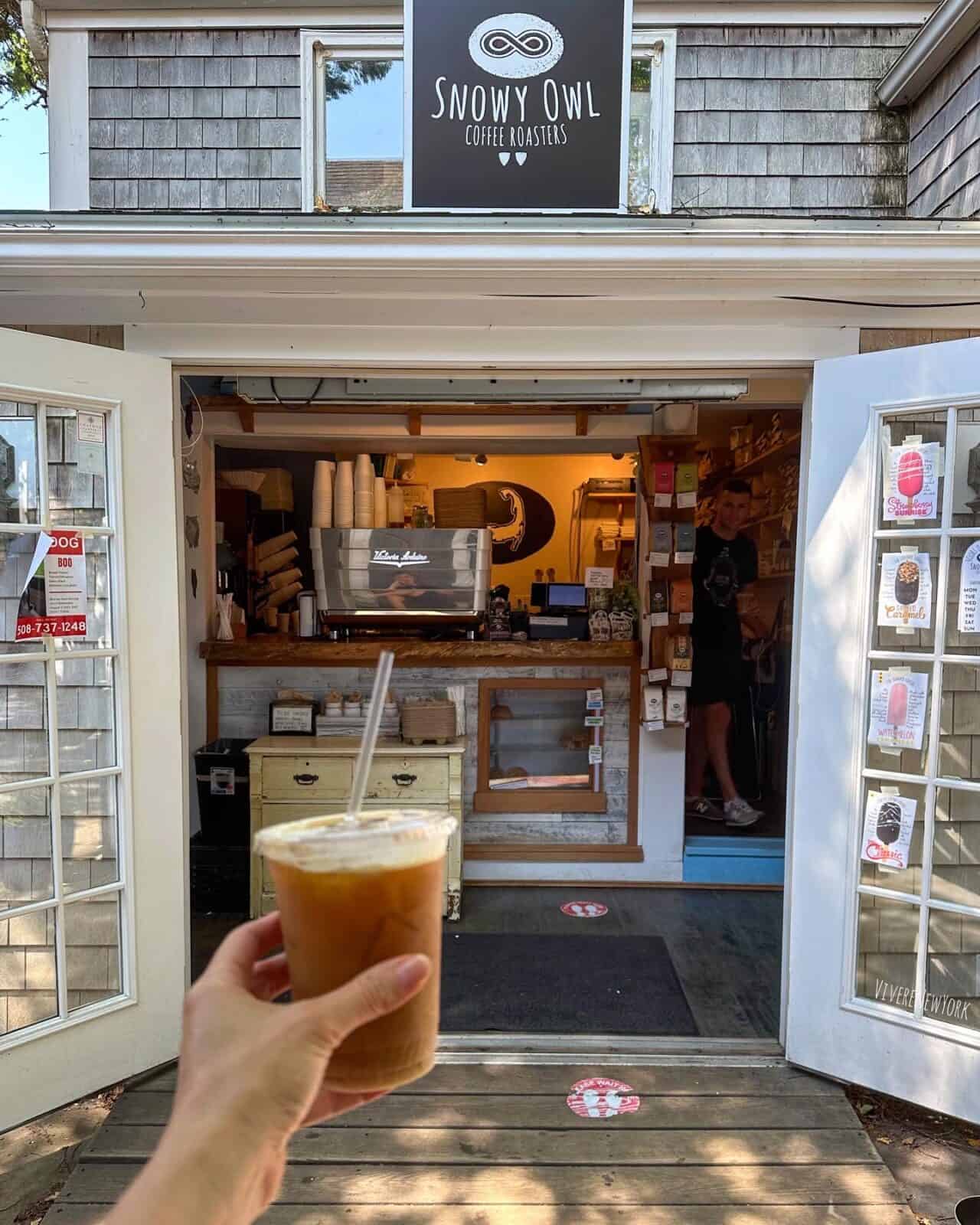 A first-person perspective holding a clear plastic cup of iced coffee with a straw in front of the white double French doors of Snowy Owl Coffee Roasters' walk-up window kiosk in Chatham, Massachusetts.