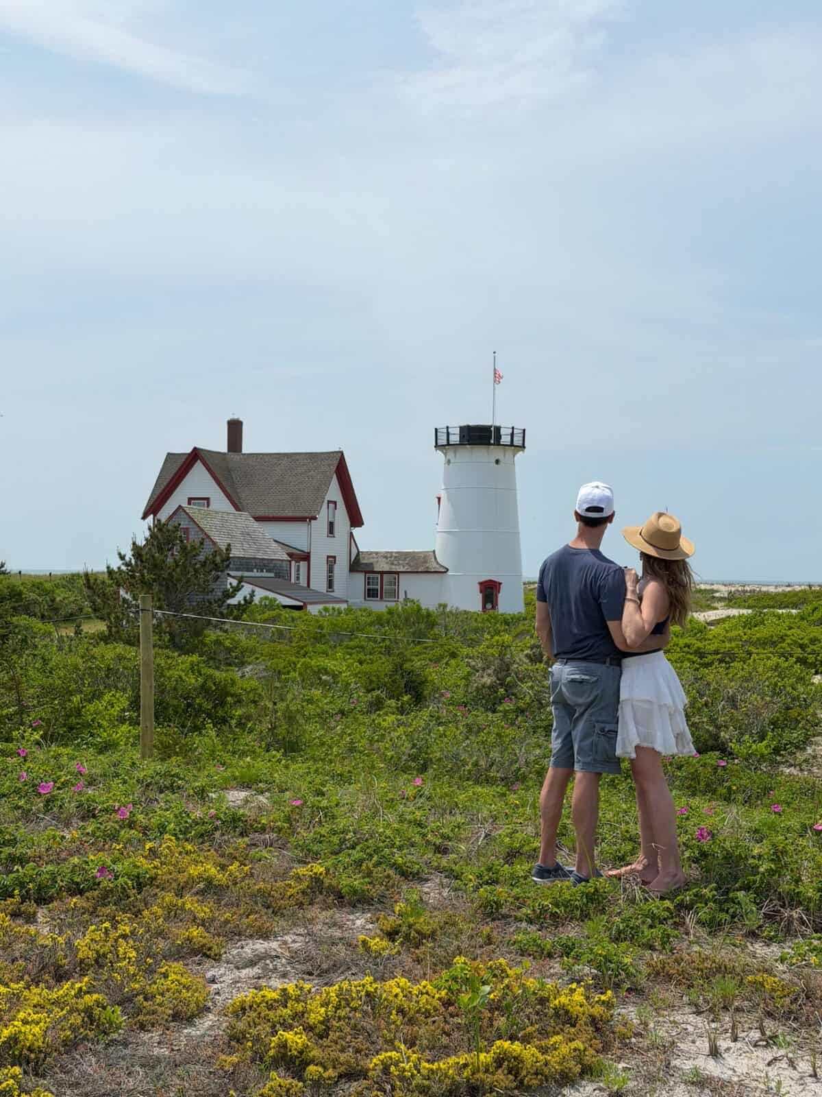 A romantic couple stands side-by-side in a field of green beach brush, bright yellow wildflowers, and pink beach roses, looking out at the white tower and red-trimmed keeper's house of Stage Harbor Lighthouse under a bright blue sky in Chatham, Massachusetts