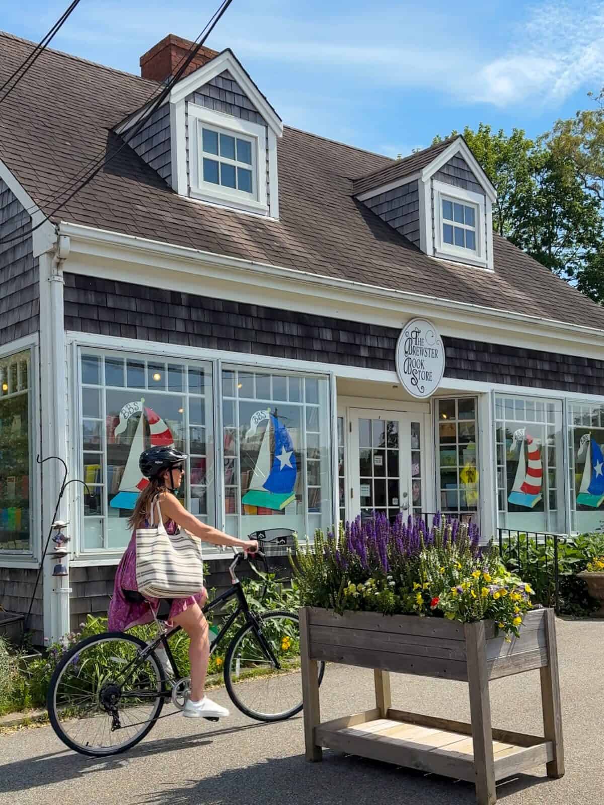 Laura wearing a helmet, pink dress, and a striped tote bag rides her black bicycle past the charming weathered-shingle exterior of The Brewster Book Store, which features large front windows decorated with sailboat illustrations.