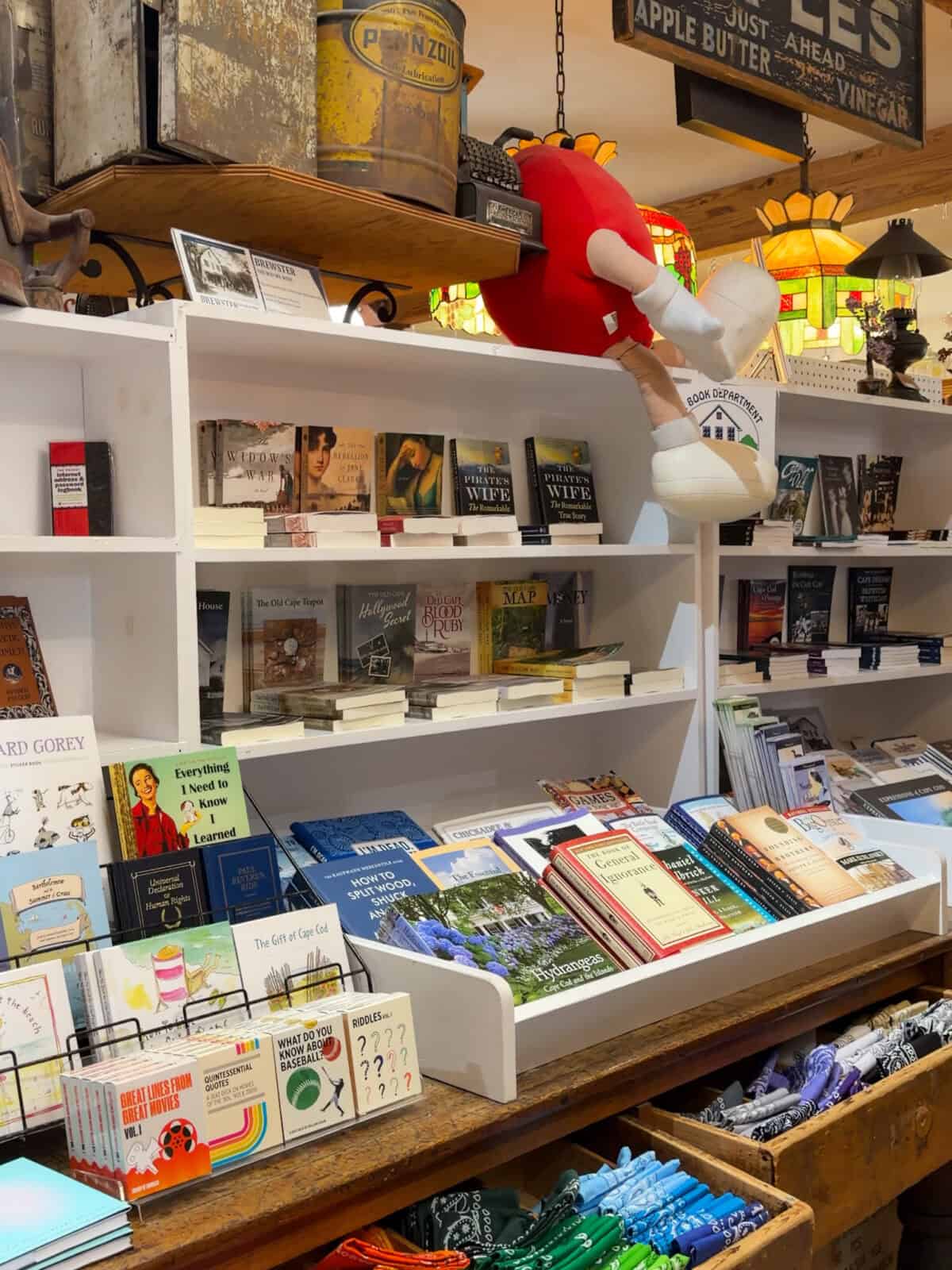 A white wooden bookshelf displaying a curated selection of regional Cape Cod history books, local guides, and literature inside The Brewster Store, accented by vintage antiques on the top shelf and a whimsical stuffed character hanging overhead.