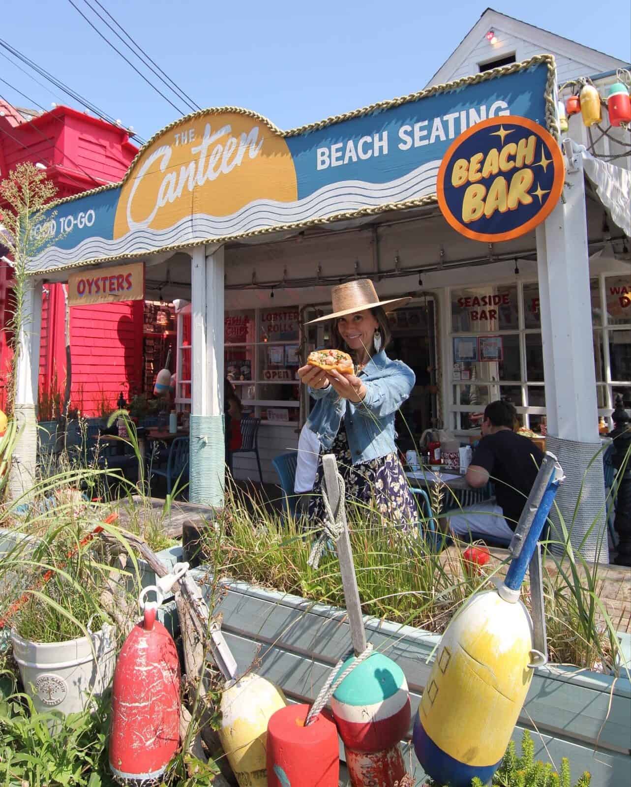 Laura in a denim jacket and straw hat smiles while presenting a fresh hot lobster roll towards the camera at the vibrant outdoor patio of The Canteen restaurant, surrounded by colorful nautical buoys and beach grasses.