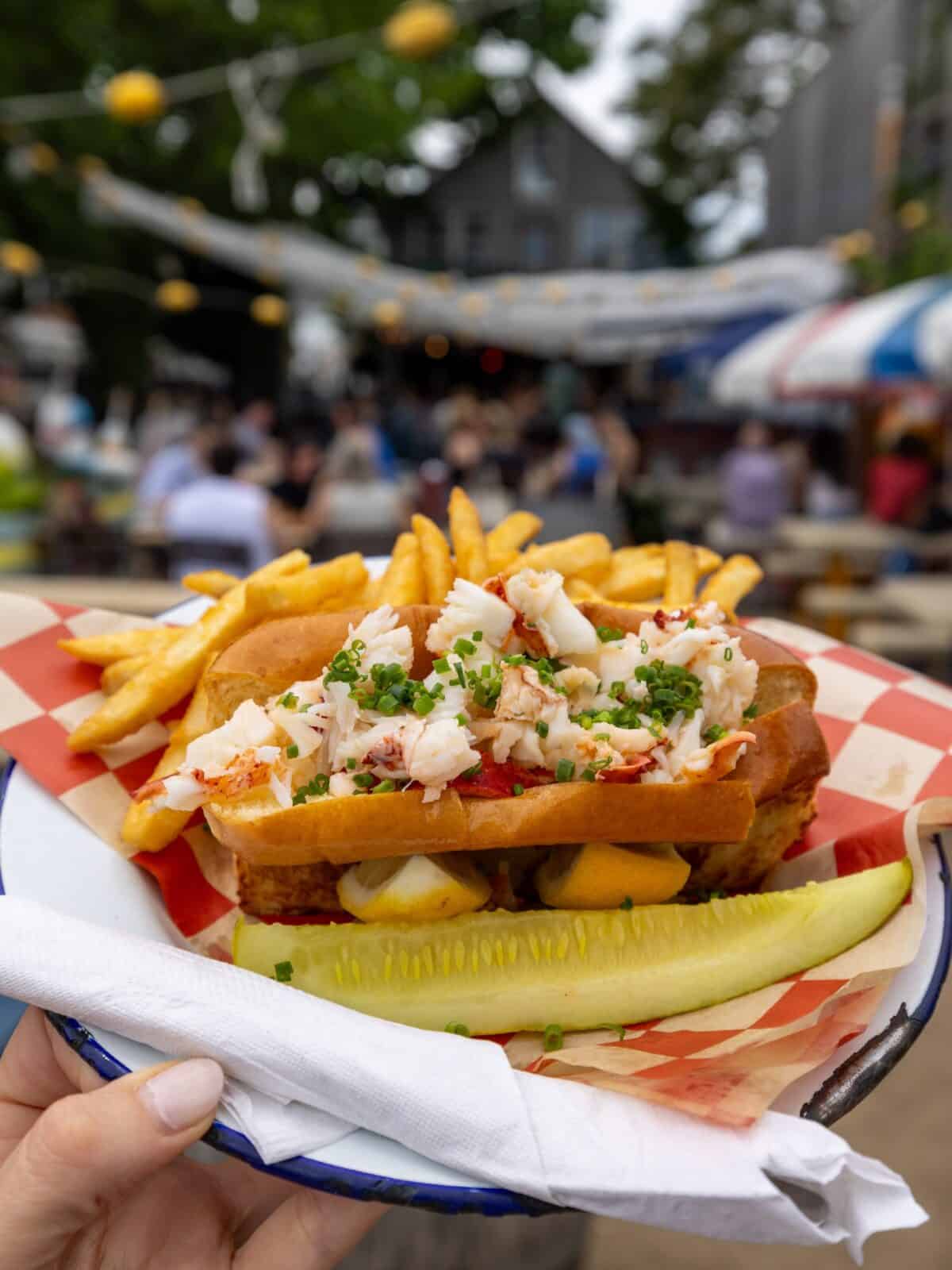 A close-up, top-down shot of a classic New England lobster roll loaded with fresh chunks of claw and tail meat, garnished with chopped chives, and served with golden french fries and a large dill pickle spear on a red-and-white checkered paper plate at The Canteen patio.