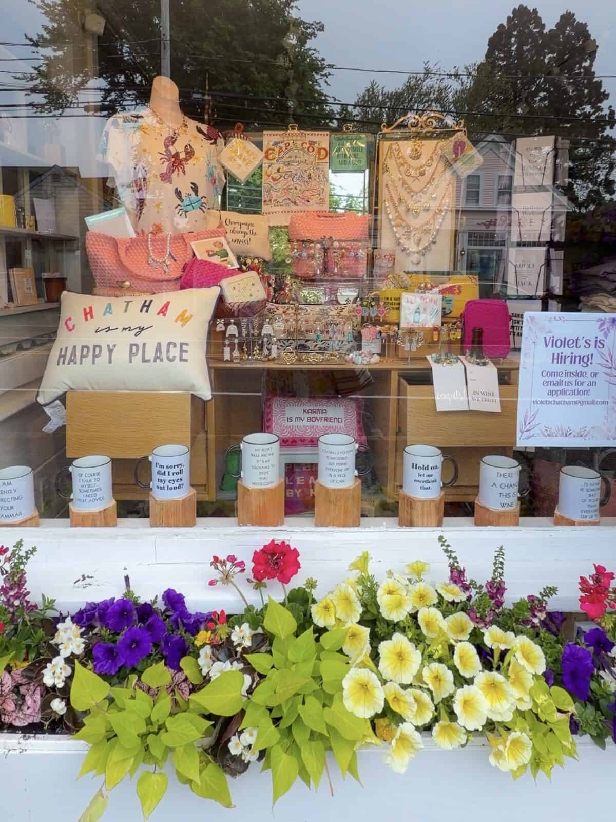 The colorful storefront window display of Violet's boutique in Chatham, showcasing local quote mugs, a "Chatham is my Happy Place" cushion, resort wear, and a prominent hiring sign above a window box of yellow and purple flowers