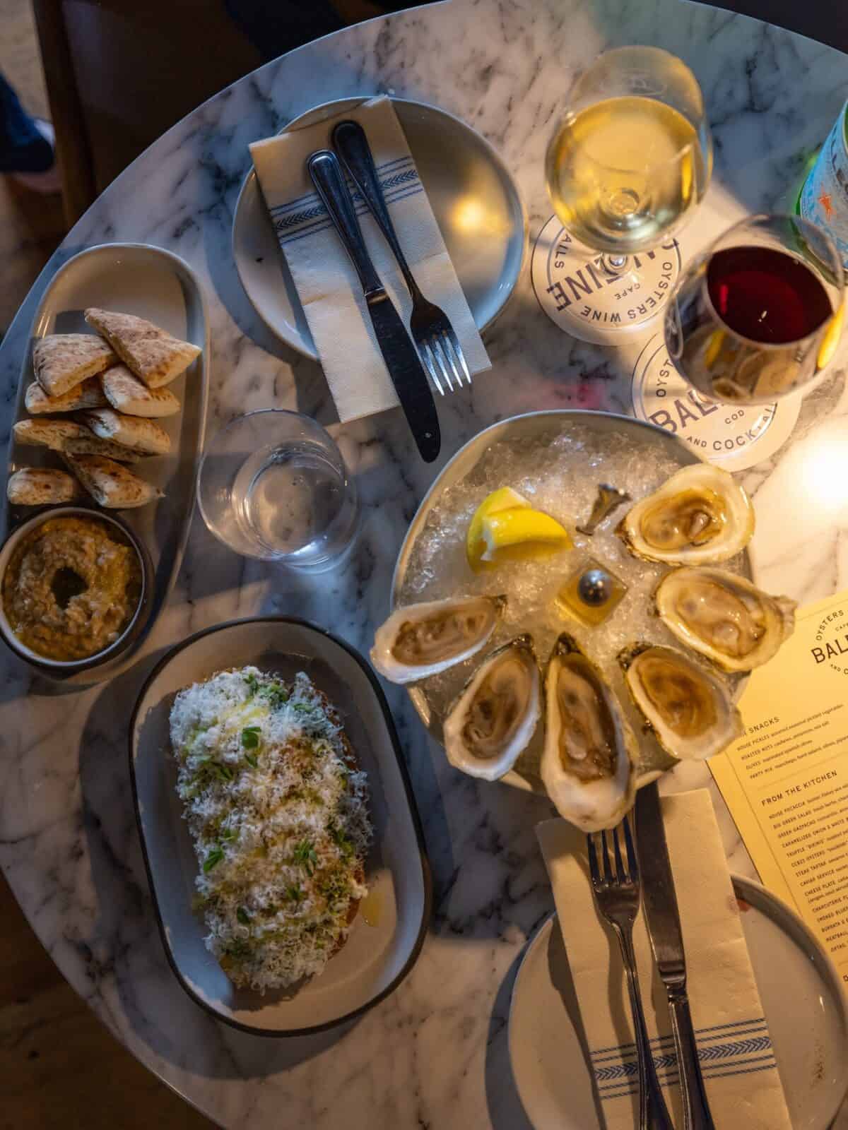 An overhead flat-lay shot of a marble restaurant table displaying a platter of six fresh raw oysters on ice with lemon, glasses of red and white wine, pita bread with dip, and an artisanal toast dish heavily topped with shaved cheese.